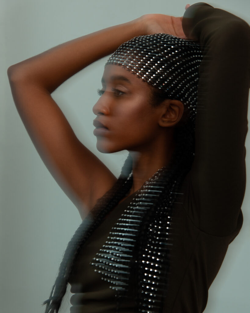 Model Ariam Tefferi of another species agency shot by Mégane Brunette, Make up by Nelly C. The model is looking to her side with both arms up to her head, wearing a bejewelled hair wrap, there is motion blur.