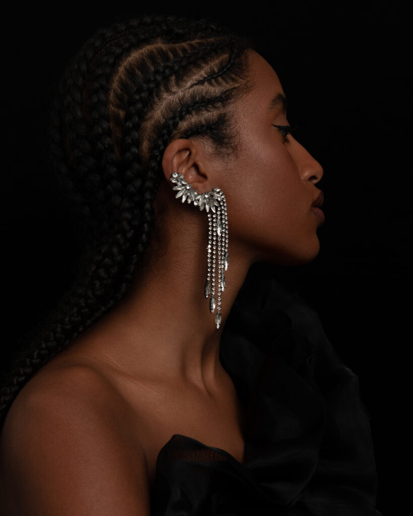 Model Ariam Tefferi of another species agency shot by Mégane Brunette, Make up by Nelly C. The model is showing of her side profile on a black backdrop and wearing a silver ear piece jewelry.