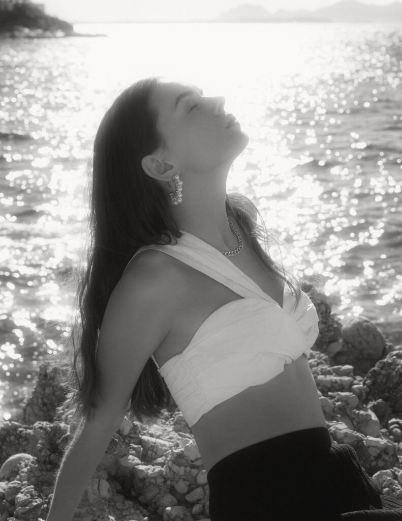 Model Elisa Bardoux of Enjoy Models Agency is sitting on rocks, leaning back. It is a sunny day on the French Riviera and the sun is reflecting on the water creating a hazy effect photographed by Mégane Brunette