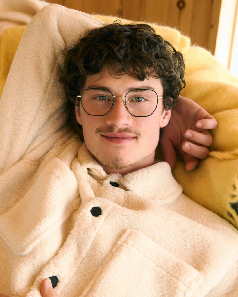 In a photoshoot for eyewear company Bonlook, model Max wearing a cozy shearling outfit, laying down on a couch. Styled by Gabriel Dupuis, hair and makeup by Genevieve Lenneville of Folio Management, photographed by Megane Brunette.